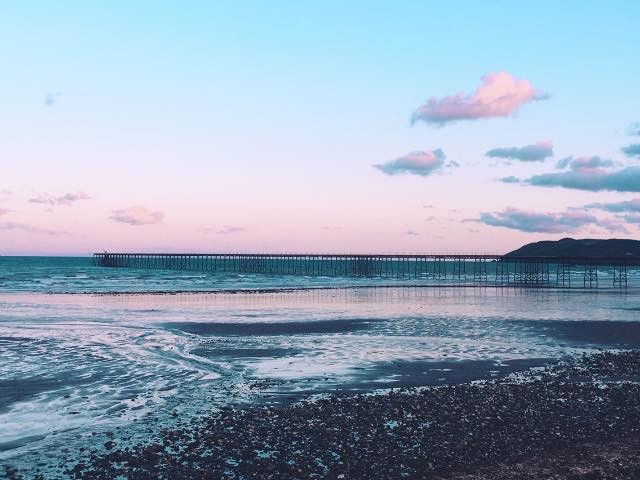 A picturesque photo of Ramsey Bay showing pink and blue clouds, a calm sea and the pier in the background. 