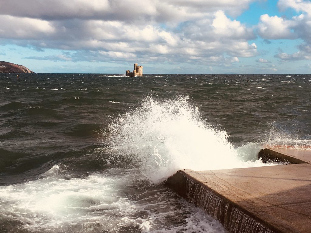 The Tower of Refuge in Douglas from the coast, with some crashing waves in the foreground on a cloudy day with some blue skies.