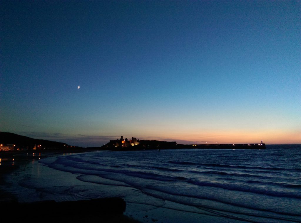 Striking Peel Castle at night in the distance, with the waves of the sea in the foreground and a bluey orange sunset 