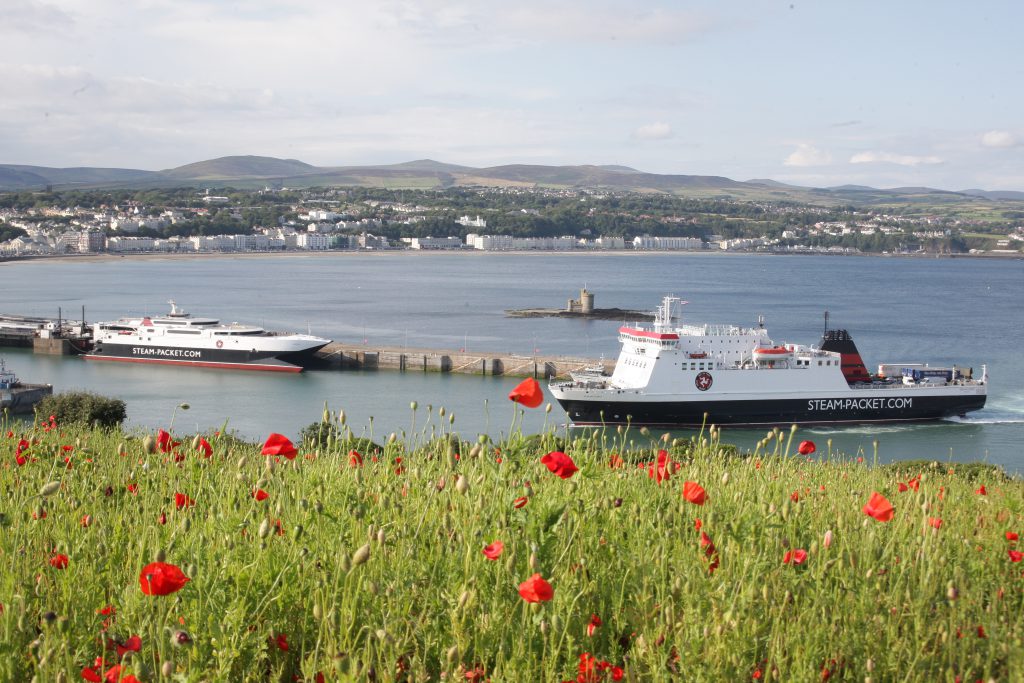 A photo taken from Douglas Head which features Steam Packet Company vessel Ben-my-Chree sailing into Douglas Harbour with Manannan already in the bay. Red poppies and green grass in the foreground. 