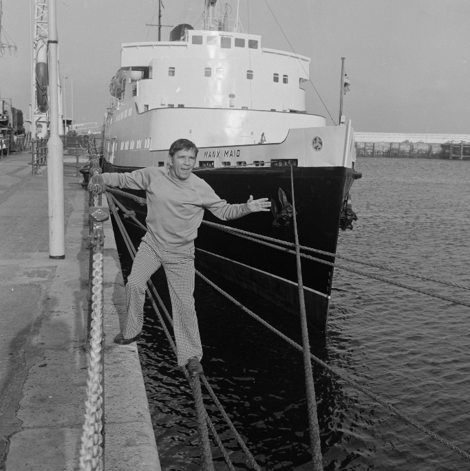 A black and white photo of Norman Wisdom posing happily next to Steam Packet Company vessel Manx Maid in Douglas Harbour in 1977