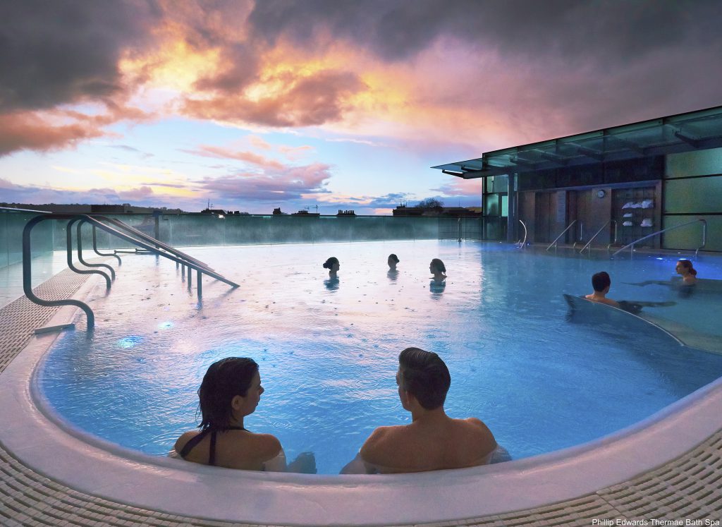 Rooftop Pool at the Thermae Bath Spa, Bath, England. Couple relaxing in the pool looking out to a pink sky. 