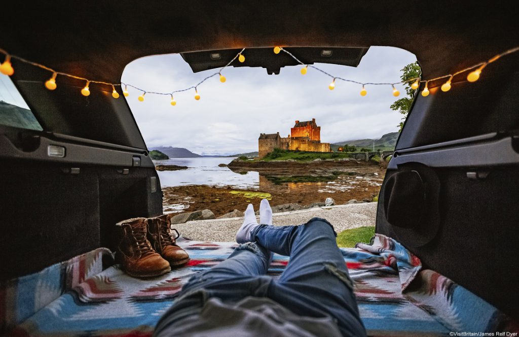 Man lying in cosy campervan with blankets and fairly lights looking at Eilean Donan, Scotland.