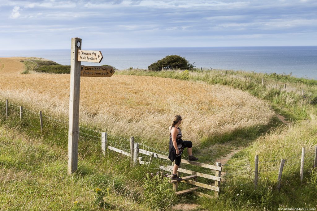 Woman climbing over turnstile, Cleveland Way, North Yorkshire, England. Views out to sea. 