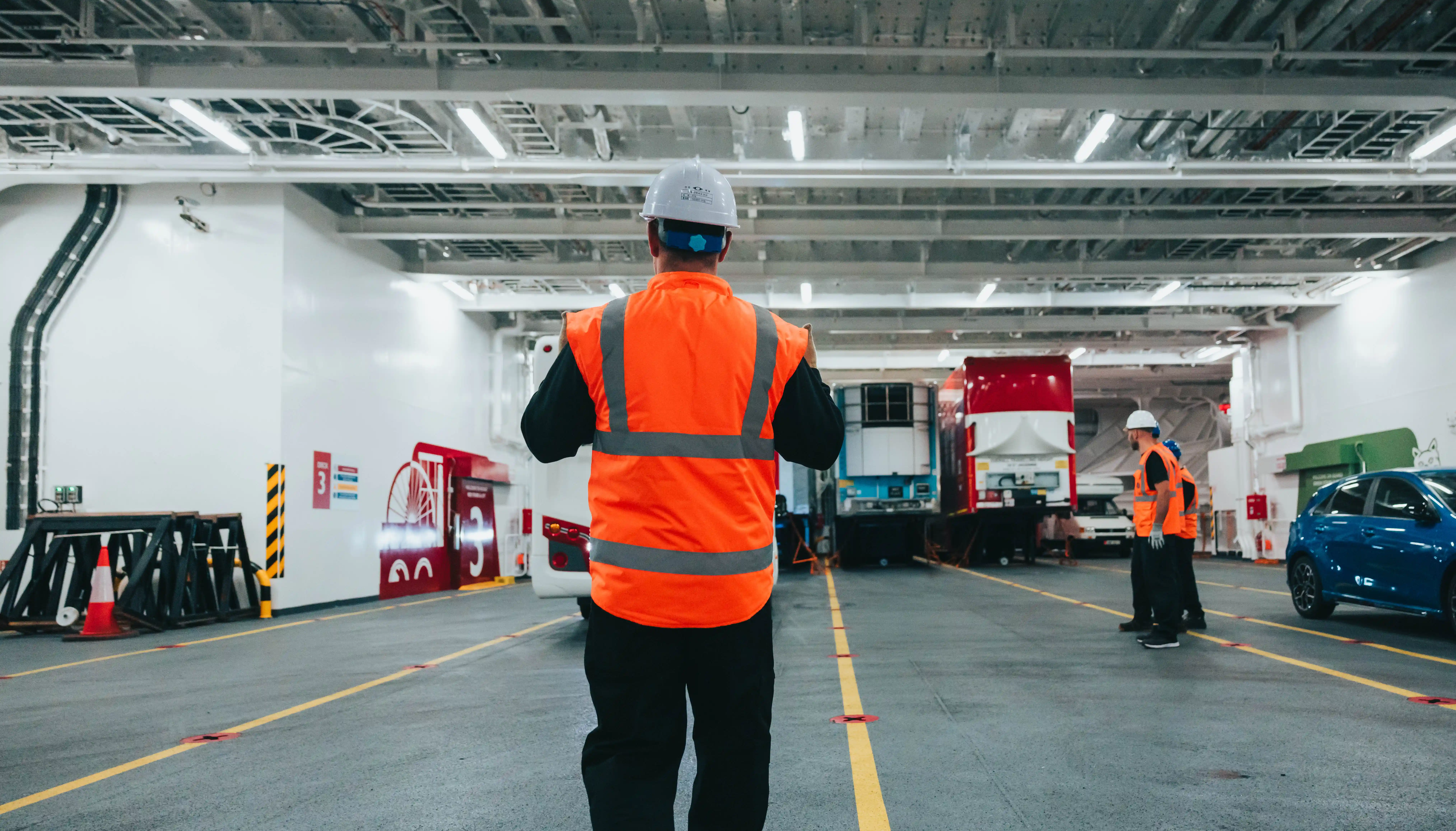 A number of Isle of Man Steam Packet crew dressed in personal protective equipment at work guiding passengers as they park vehicles on Manxman's deck 3. In the background a number of freight trailers are parked.