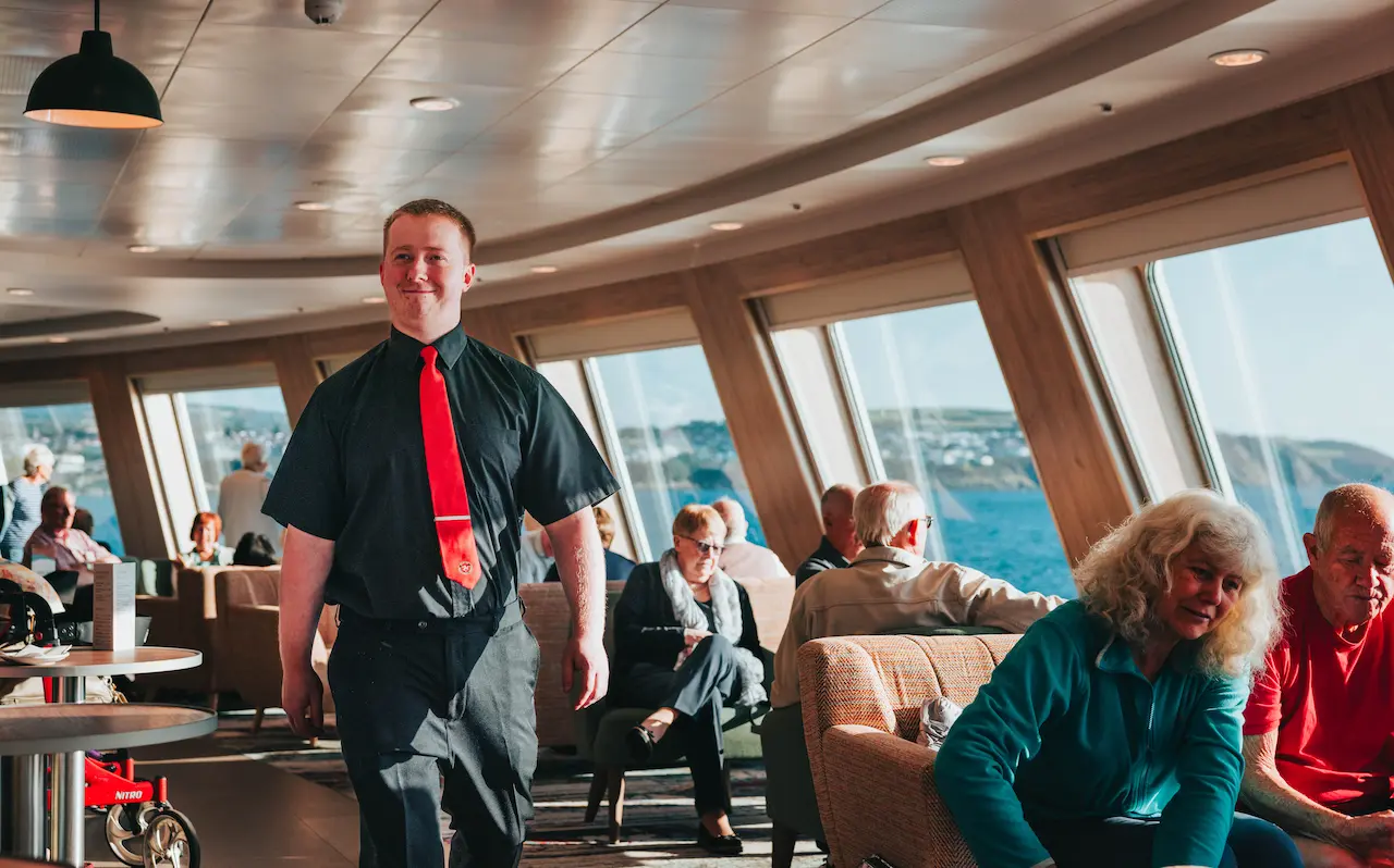 A Passenger Services Assistant walks between passengers seated at sofas and armchairs in the forward lounge of Manxman