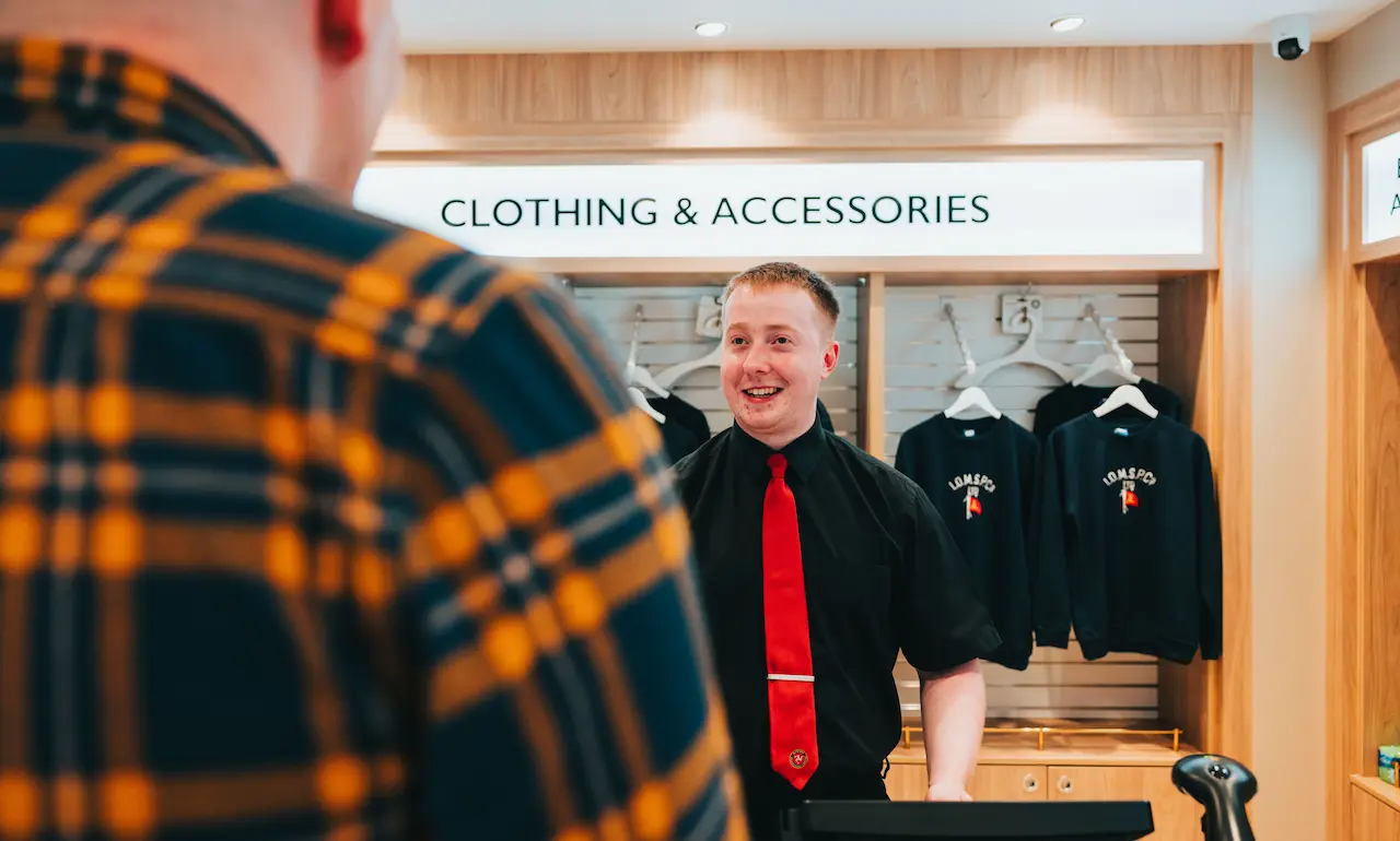 A Passenger Services Assistant aboard Manxman serves a customer in the vessel's shop. In the background IOMSPC jumpers hang on a product rail